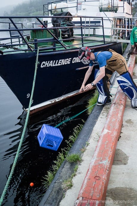 Boy wearing hockey padding throwing milk carton over edge of wharf to get hockey ball from the water. JHHG3-1000647
